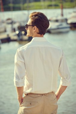 Denizde yaz tatili. Beyaz gömlek, şort ve siyah güneş gözlüğü takan bir adam deniz suyunun yanındaki yat limanında duruyor. Zarif erkek modası. 