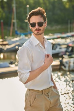 Denizde yaz tatili. Beyaz gömlek, şort ve siyah güneş gözlüğü takan bir adam deniz suyunun yanındaki yat limanında duruyor. Zarif erkek modası. 