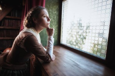 Romanın kadın kahramanı. 18. yüzyıl İskoç elbisesi giymiş güzel bir aristokrat kız klasik bir iç mimariye sahip bir odada düşünceli bir şekilde pencereden dışarı bakar. 18. yüzyıl tarzı.