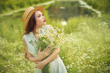 Yazın hasır şapkalı, kahverengi saçlı romantik bir kız bir buket taze papatya çiçeği ve gülümsemeyle bir yaz günü dışarıda duruyor. Yaz güzelliği, makyaj ve çiçekler. Boşluğu kopyala.