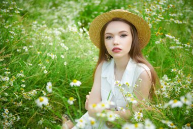 Yaz güzelliği, makyaj ve çiçekler. Yazlık hasır şapkalı güzel kahverengi saçlı bir kız yeşil çimenlerin ve papatyaların arasında oturuyor.. 