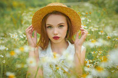 Hasır şapkalı hassas bir kız yeşil çimenler ve papatyalarla çevrili bir şekilde tarlada oturuyor. Yaz güzelliği, makyaj ve çiçekler..