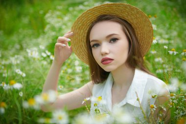 Hasır şapkalı hassas bir kız yeşil çimenler ve papatyalarla çevrili bir şekilde tarlada oturuyor. Yaz güzelliği, makyaj ve çiçekler..