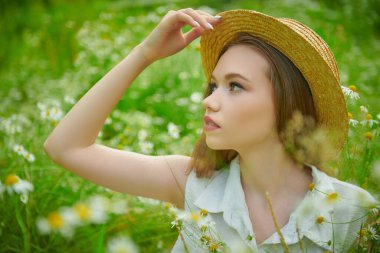 Yazlık hasır şapkalı güzel kahverengi saçlı bir kız yeşil çimenlerin ve papatyaların arasında oturuyor. Yaz güzelliği, makyaj ve çiçekler..