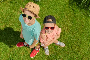 Güneşli bir yaz gününde yeşil bir parkta gülümseyen ve ayakta duran yaz kıyafetleri ve güneş gözlükleriyle bir erkek ve kız kardeşinin portresi. Mutlu yaz tatilleri. Mutlu aktif çocuklar.