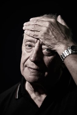 70 'li yaşlarda, elini başının üstüne koyup sakin bir gülümsemeyle kameraya bakan yaşlı bir adamı yakından çek. Siyah bir arkaplanda stüdyo resmi.
