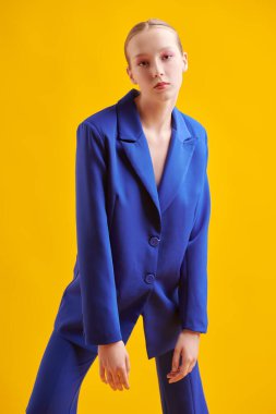 Yumuşak pembe makyajlı güzel bir manken sarı stüdyo arka planında zarif mavi bir takım elbiseyle poz veriyor. Parlak yaz renkleri. Moda ve güzellik.  