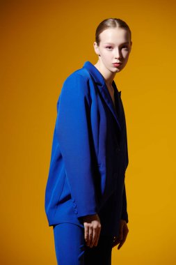 Parlak yaz renkleri. Yumuşak pembe makyajlı güzel bir manken sarı stüdyo arka planında zarif mavi bir takım elbiseyle poz veriyor. Moda ve güzellik.