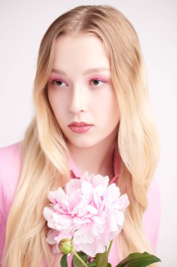 Makyaj ve makyaj malzemeleri. Narin pembe makyajlı güzel sarışın bir kızın portresi. Şakayık çiçeğiyle poz veriyor. Beyaz arka plan. Parfüm. Kadın güzelliği.