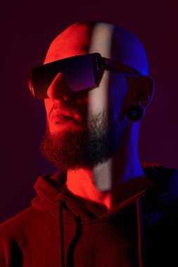 Dudak delen, güneş gözlüğü takan ve siyah kapüşonlu sakallı vahşi bir adamın portresi. Stüdyo çekimleri siyah arkaplan ile karışık koyu kırmızı ve mavi ışıkta.