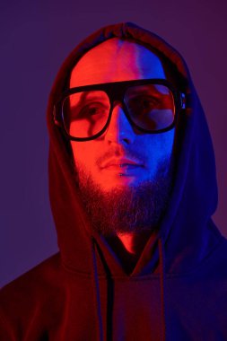 Kel, sakallı, dudağında piercing olan, siyah kapüşonlu ve büyük gözlüklü vahşi bir adamın karışık renkli portresi..