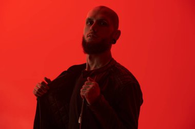 Dudaklarında ve kulaklarında piercingler olan vahşi, sakallı bir adam kırmızı stüdyo arka planında siyah deri ceketli poz veriyor..