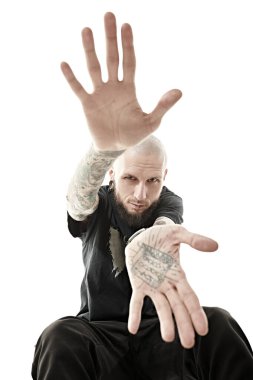 Vücudunda dövmeleri olan, kulaklarında ve dudaklarında piercingler olan, siyah elbiseler giymiş, avuçlarını kameraya uzatan kel bir hippi portresi. Beyaz bir arkaplanda stüdyo çekimi.