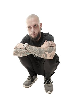 Kafası kazınmış, sakallı, vücudunda dövmeleri olan, siyah kıyafetler giymiş, çömelmiş ve kameraya dikkatlice bakan gayri resmi bir vahşi adam. Stüdyo beyaz arkaplan.