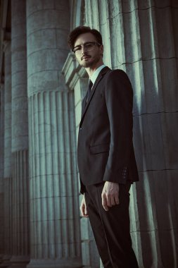 Beyaz gömlekli ve kravatlı, klasik siyah takım elbiseli yakışıklı genç bir adam bir caddedeki sütunlarda poz veriyor. İş adamı. Erkek modası.