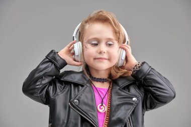 Parlak makyajlı, modaya uygun kıyafetleri ve aksesuarları olan sevimli bir kız ve rock müzisyeni kulaklıkla müzik dinliyor. Gri stüdyo geçmişi. Çocuk modası. DJ çocuk.