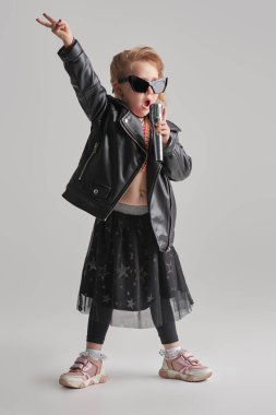 Havalı küçük kız - Rock yıldızı şarkı söylüyor ve dans ediyor. Stüdyonun arka planına karşı tam boy portre. Çocuklar için rock and pop müzik kültürü. Çocuk modası.