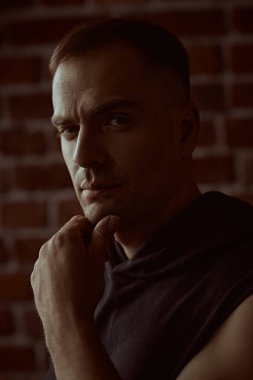 Siyah kapüşonlu güçlü bir adamın yakın plan portresi, odaklanmış ve yoğun bir şekilde kameraya bakıyor. Arka planda tuğla duvar var. Halk konsepti. Erkekler, erkeklik ve güç..