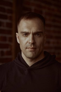 Erkekler, erkeklik ve güç. Siyah kapüşonlu güçlü bir adamın yakın plan portresi, odaklanmış ve yoğun bir şekilde kameraya bakıyor. Arka planda tuğla duvar var. Halk konsepti. 