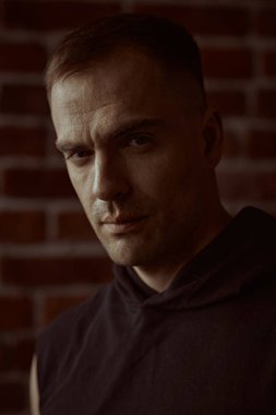 Siyah kapüşonlu güçlü bir adamın yakın plan portresi, odaklanmış ve yoğun bir şekilde kameraya bakıyor. Arka planda tuğla duvar var. Halk konsepti. Erkekler, erkeklik ve güç..