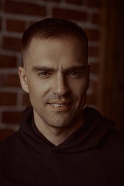 Siyah kapüşonlu gülümseyen bir adamın yakın plan portresi. Kameraya bakıyor. Arka planda tuğla duvar var. Halk konsepti. Erkekler, erkeklik ve güç..