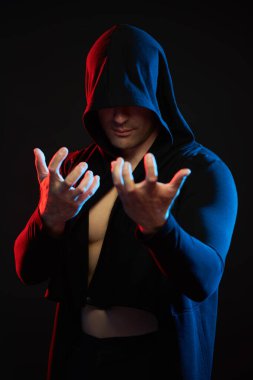 Güçlü, atletik bir adam duruyor, yüzünü bir kaputun altına gizliyor, gergin kollarını kameraya doğru açıyor. Stüdyo portresi ve karışık ışık. Güç sporları. Boksör. Savaşçı..