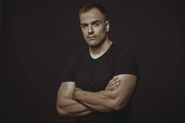 Atletik yapılı yakışıklı ve güçlü bir adam sakin ve emin bir şekilde duruyor. Kolları göğsünün üzerinde çapraz duruyor. Metin için alanı olan siyah arkaplan. İnsanlar ve duygular. Aktif, sağlıklı bir yaşam tarzı. Spor..