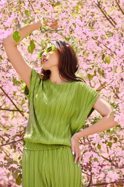 Bahar güzeli. Parlak yeşil elbiseli ve limon bardaklı güzel bir Asyalı kızın portresi çiçek açan sakura çiçeklerinin arasında duruyordu. Mutlu bahar günleri. Bahar ve yaz modası.