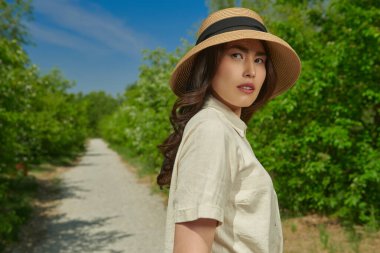 Güzel bir Asyalı genç kadın yemyeşil ve mavi gökyüzünün zeminine karşı bir yaz parkının arka sokağında yürüyor. Mutlu güneşli günler. Yaz tatili.