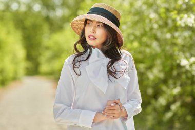 Şapkalı güzel bir Asyalı genç kadın yemyeşil bir arka planda yaz parkında yürüyor. Mutlu güneşli günler. Yaz tatili.