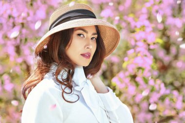 Bahar çiçekleri ve güzellik. Beyaz bluz ve hasır şapkalı Asyalı romantik bir genç kadının portresi çiçek açan sakura çiçeklerinin arasında duruyordu..