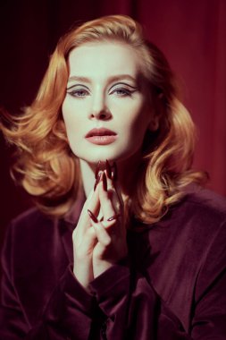 Pahalı kadife ceketli çekici kızıl saçlı bir kadının portresi kırmızı kadife perdelerin arka planında. Lüks ve cazibe tarzı. Kadın Güzelliği ve Moda. Film ve tiyatro oyuncusu.