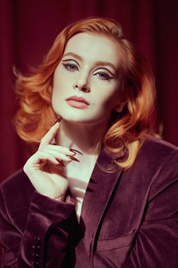 Moda ve kadınlık. Pahalı kadife ceketli, loş ışıklı bir odada kızıl saçlı çekici bir genç kadın. Kadın güzelliği ve tarzı. Oklu zarif bir makyaj. Zarif saç stili..