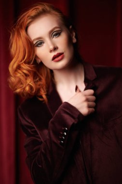 Şık saç stili. Pahalı kadife ceketli, loş ışıklı bir odada kızıl saçlı çekici bir genç kadın. Moda ve kadınlık. Kadın güzelliği ve tarzı. Oklu zarif makyaj..