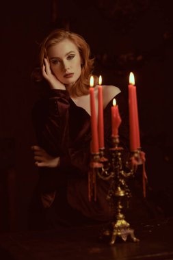 Pahalı kadife ceketli çekici kızıl saçlı genç bir kadın mumların arkasındaki kırmızı kadife perdelerin arkasında. Zarif saç stili ve oklu makyaj. Kadın Güzelliği ve Moda.