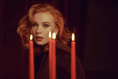 Pahalı kadife ceketli çekici kızıl saçlı genç bir kadın mumların arkasındaki kırmızı kadife perdelerin arkasında. Zarif saç stili ve oklu makyaj. Kadın Güzelliği ve Moda.