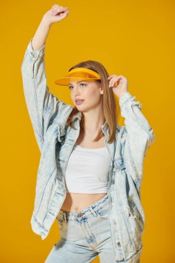 Denim gençlik tarzı. Kot elbiseli güzel sarışın bir kızın portresi ve sarı stüdyo arka planında şık sarı bir şapka. Yaz modası. Spor kıyafetleri..