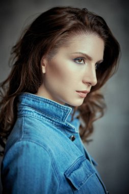 Gri bir arka planda kot gömlekle poz veren güzel kahverengi dalgalı saçlı çekici bir kızın portresi. Güzellik ve güven. Denim biçimi.