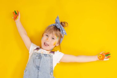 Çocuklar için yaratıcılık ve çizim. Küçük sanatçı. Renkli boyalarla boyanmış beş yaşında tatlı bir kız, sarı arka planda yerde mutlu bir şekilde yatıyor. Çocukların hayalleri ve hayalleri.