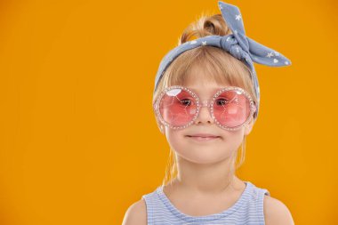 Çocuk modası. Küçük moda kızı. Stüdyonun sarı arka planında poster giymiş beş yaşında tatlı bir kızın portresi. Gözlük stili.