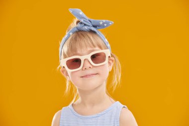 Küçük moda kızı. İlan tarzı giyinmiş 5 yaşında tatlı bir kız kameraya bakıp gülümsüyor. Stüdyo sarı arka plan ve fotokopi alanı. Çocuk modası. Gözlük stili.