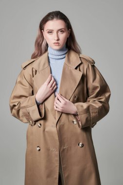 Gri stüdyo arka planında bej renkli bir trençkotla poz veren çekici kendine güvenen kız model. Kadın modası. Sonbahar-ilkbahar.