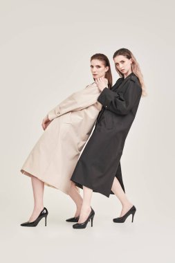 Beyaz bir stüdyo arka planında modaya uygun paltolarla poz veren kendine güvenen iki kadın modelin tam boy çekimi. Yarı sezon modası. 