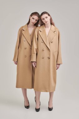 Uzun bej keçeli ceketlerle poz veren iki güzel manken kızın tam boy portresi. Beyaz stüdyo geçmişi. Yarı sezon modası.