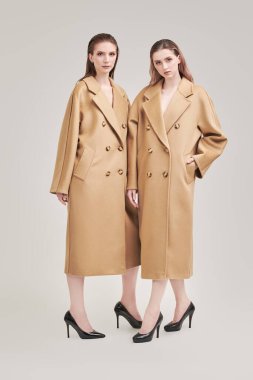 Yarı sezon modası. Uzun bej keçeli ceketlerle poz veren iki güzel manken kızın tam boy portresi. Beyaz stüdyo geçmişi.