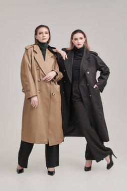 Beyaz bir stüdyo arka planında modaya uygun paltolarla poz veren kendine güvenen iki kadın modelin tam boy çekimi. Yarı sezon modası. 