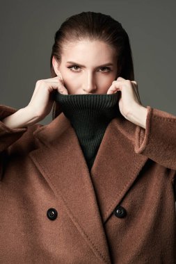 Siyah boğazlı kazak ve kahverengi keçeli palto giyen kendine güvenen şık bir kız. Gri bir arka planda stüdyo resmi. Moda fotoğrafı..