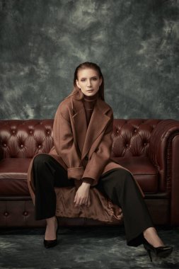 Çekici, kendine güvenen bir kadın deri koltukta poz veriyor moda keçeli ceketi, siyah pantolonu ve yüksek topuklu ayakkabıları var. Kadın modası.