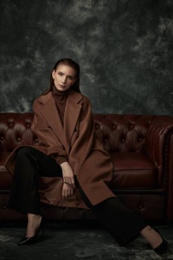 Çekici, kendine güvenen bir kadın deri koltukta poz veriyor moda keçeli ceketi, siyah pantolonu ve yüksek topuklu ayakkabıları var. Kadın modası.