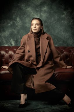 Çekici, kendine güvenen bir kadın deri koltukta poz veriyor moda keçeli ceketi, siyah pantolonu ve yüksek topuklu ayakkabıları var. Kadın modası.